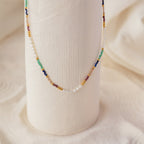 The Oasis Rainbow Beaded Necklace, featuring small pearls and colorful beads, drapes elegantly over a textured white vase—making it the perfect boho jewelry or summer accessory.