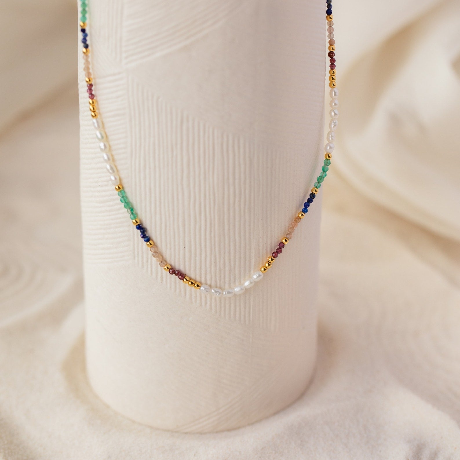 The Oasis Rainbow Beaded Necklace, featuring small pearls and colorful beads, drapes elegantly over a textured white vase—making it the perfect boho jewelry or summer accessory.