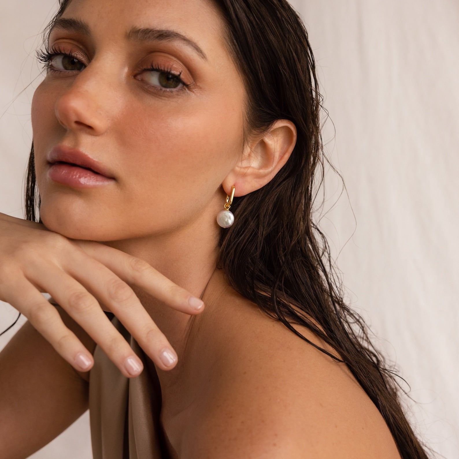 A woman with wet hair and natural makeup wears the Bridget Pearl Hoops, resting her chin on her hand.