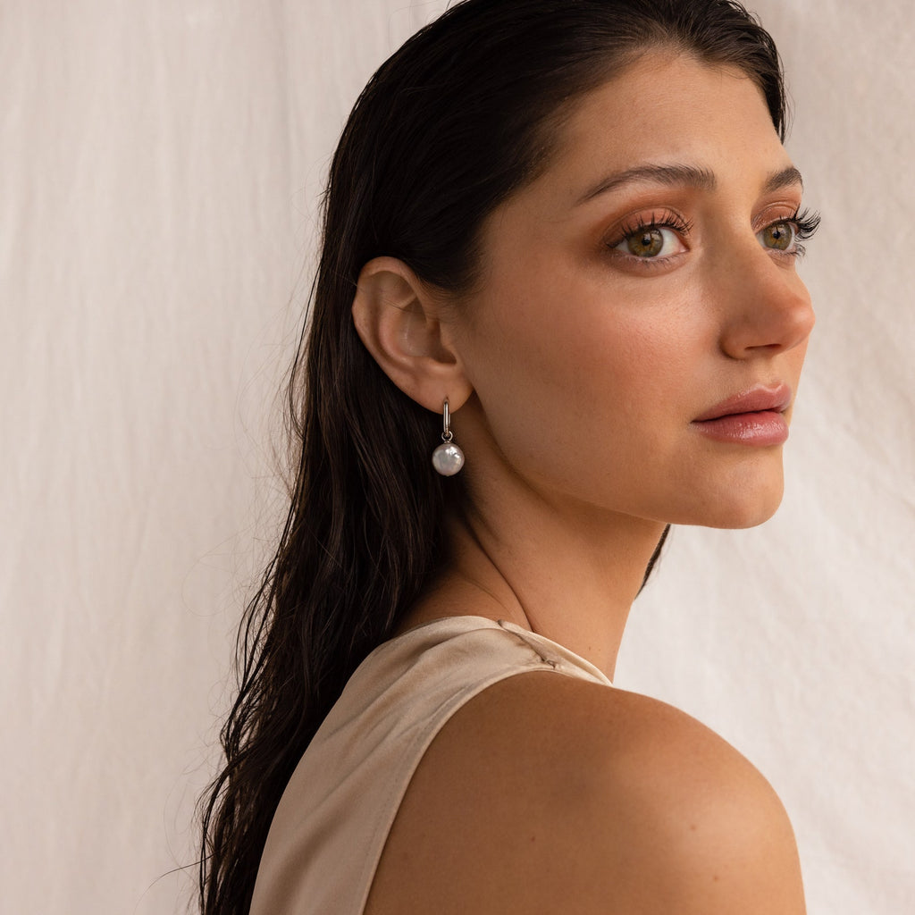 A woman with long brown hair, wearing a sleeveless beige top and elegant Bridget Pearl Hoops, gazes to the right.