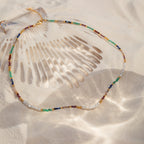 The Oasis Rainbow Beaded Necklace, an ideal summer accessory, rests on the sand next to a large seashell as sunlight casts playful shadows.
