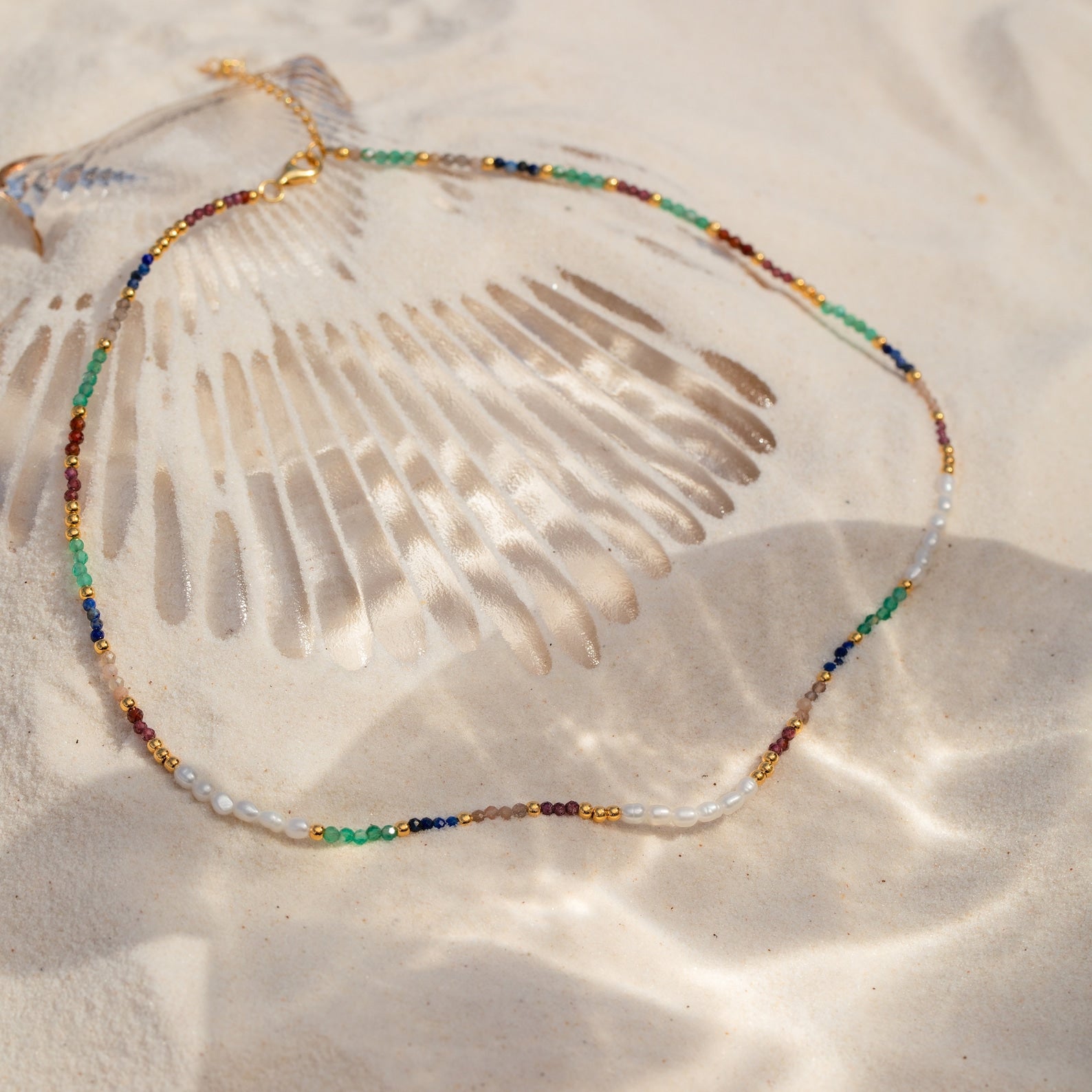 The Oasis Rainbow Beaded Necklace, an ideal summer accessory, rests on the sand next to a large seashell as sunlight casts playful shadows.