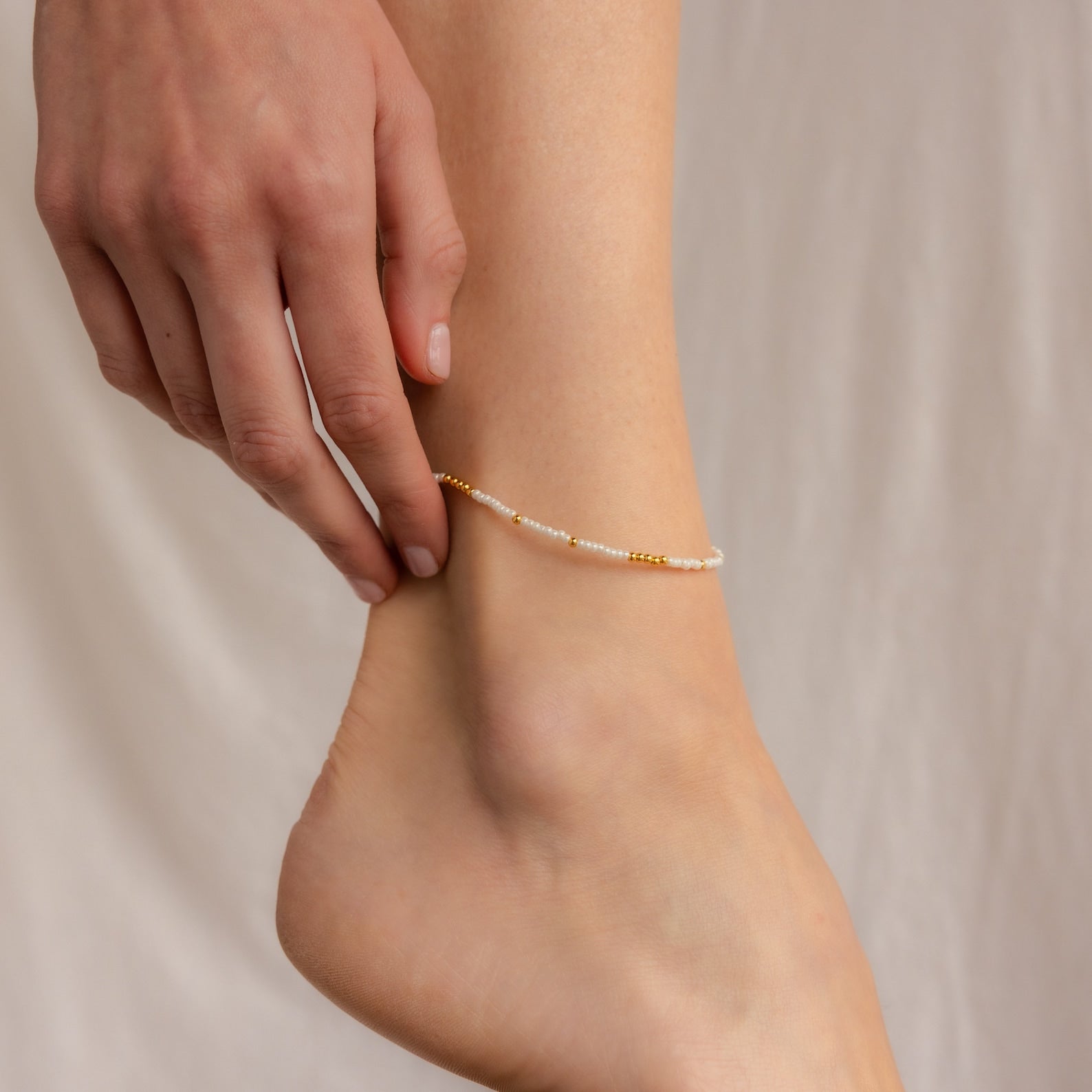 A hand touches an ankle adorned with the Dainty Pearl Beaded Anklet, featuring white and gold beads that shimmer in a delicate, beaded design.