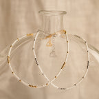 Two delicate necklaces with white, gold, and silver beads drape over a clear glass vase, perfectly complementing the Dainty Pearl Beaded Anklet for an elegant touch.
