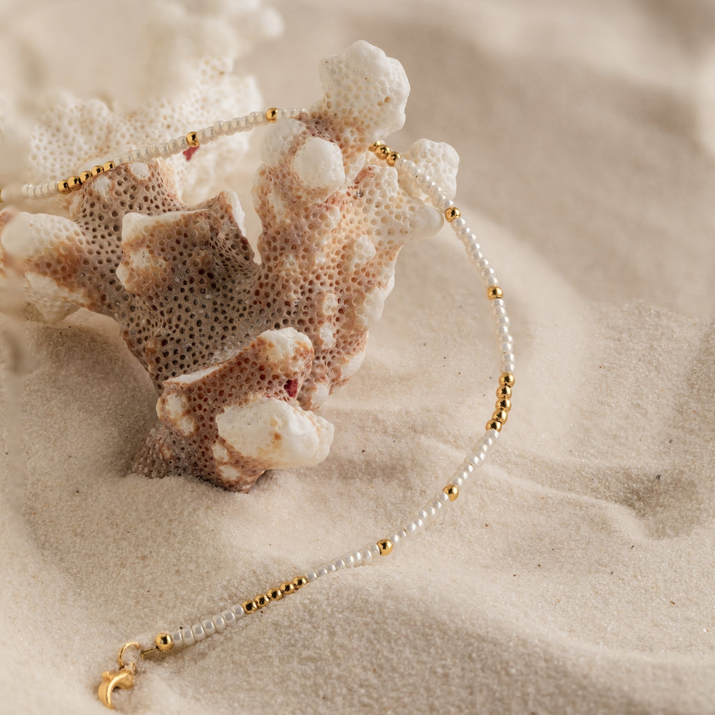 The Dainty Pearl Beaded Anklet, featuring elegant beads and gold accents, is draped over coral on soft sand.