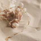 The Dainty Pearl Beaded Anklet, featuring elegant beads and gold accents, is draped over coral on soft sand.