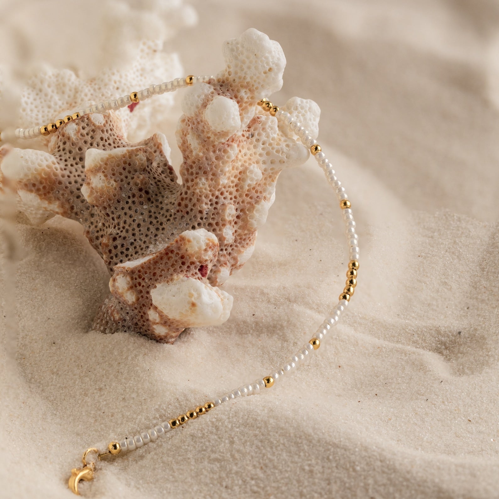 The Dainty Pearl Beaded Anklet, featuring elegant beads and gold accents, is draped over coral on soft sand.
