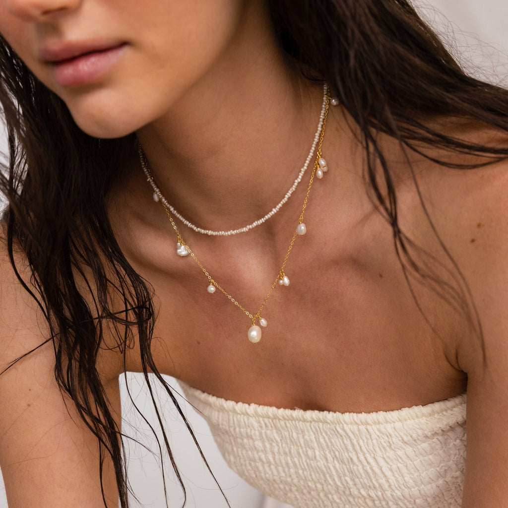 A woman layers a Baroque Pearl Station Necklace in Sterling Silver with other pearl strands, complementing her white textured strapless top and loose wavy dark hair for an elegant wedding jewelry look.