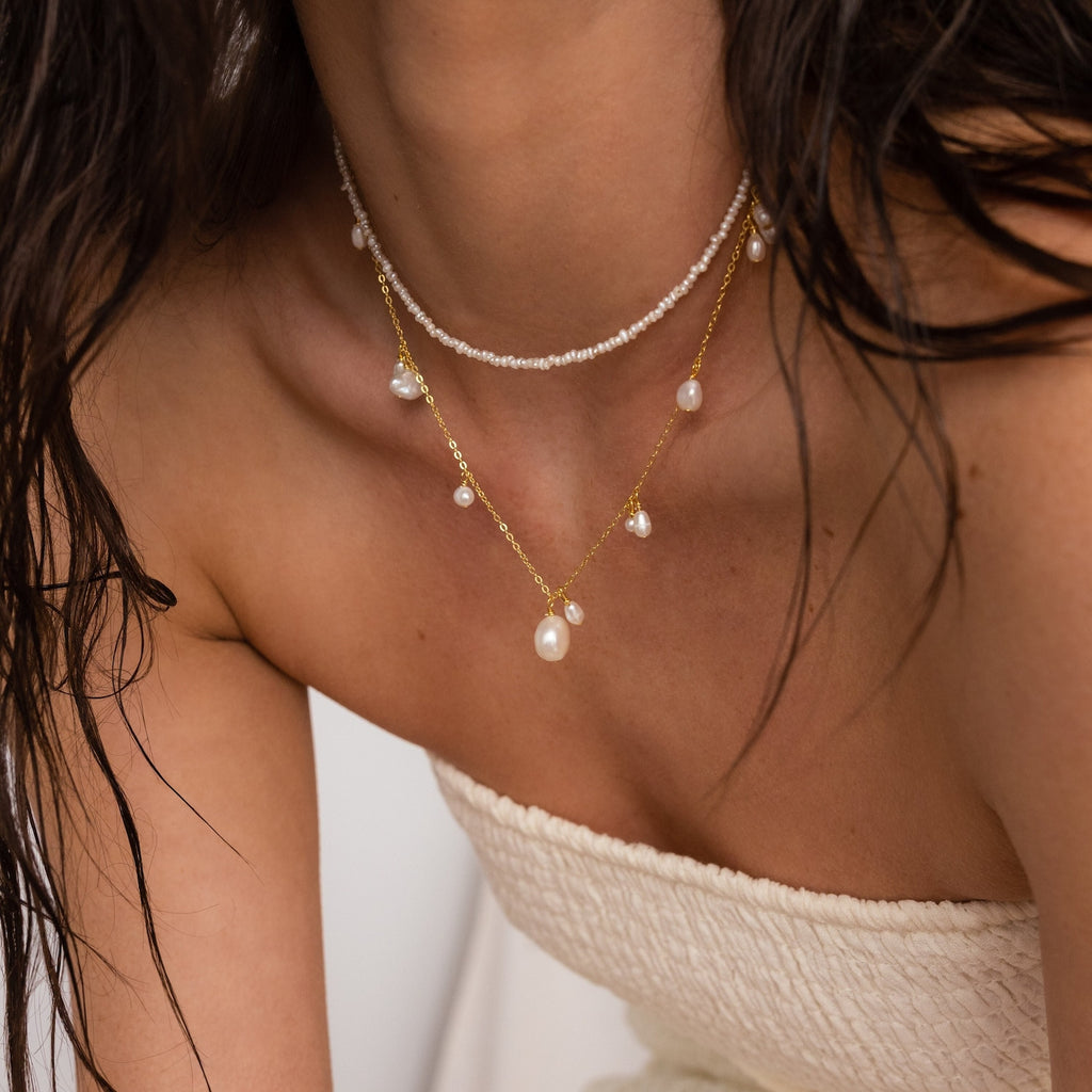 Layered necklace set with a short beaded choker and a longer gold chain adorned with dangling white pearls, worn with a strapless textured cream top.