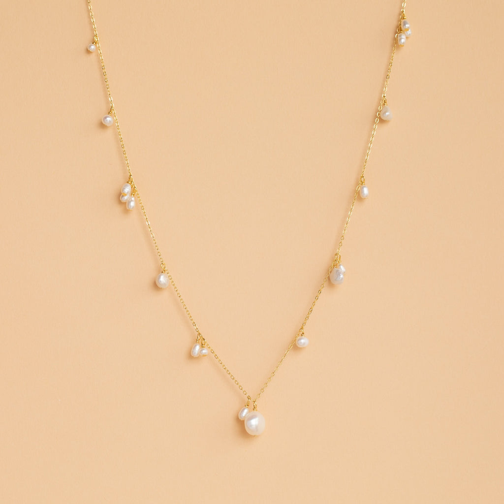 The Baroque Pearl Station Necklace in 18K Gold features a gold chain with spaced white pearls, displayed on a light beige background—an elegant choice for wedding jewelry.