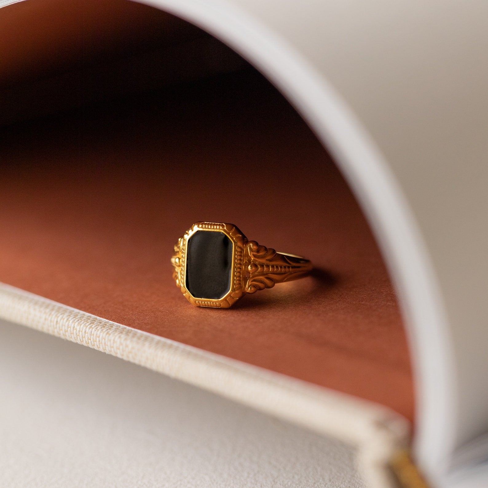 The Vintage Black Signet Ring features a chunky gold band with a black rectangular stone, displayed on a brown and beige curved surface for timeless style.