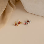 Two pairs of stud earrings, featuring the Garnet Heart Studs with red and clear stones, are displayed on a beige surface next to soft fabric and a white object.