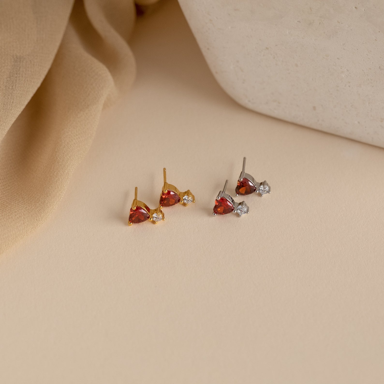 Two pairs of stud earrings, featuring the Garnet Heart Studs with red and clear stones, are displayed on a beige surface next to soft fabric and a white object.