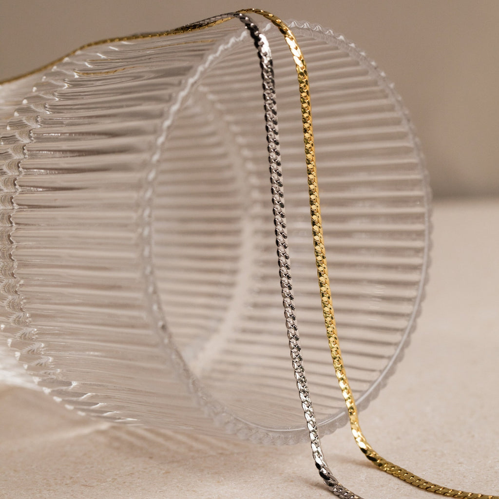 A silver Men's Snake Chain Necklace and a minimalist gold necklace rest on a ribbed clear glass set on a beige surface—stylish essentials for any men's jewelry collection.