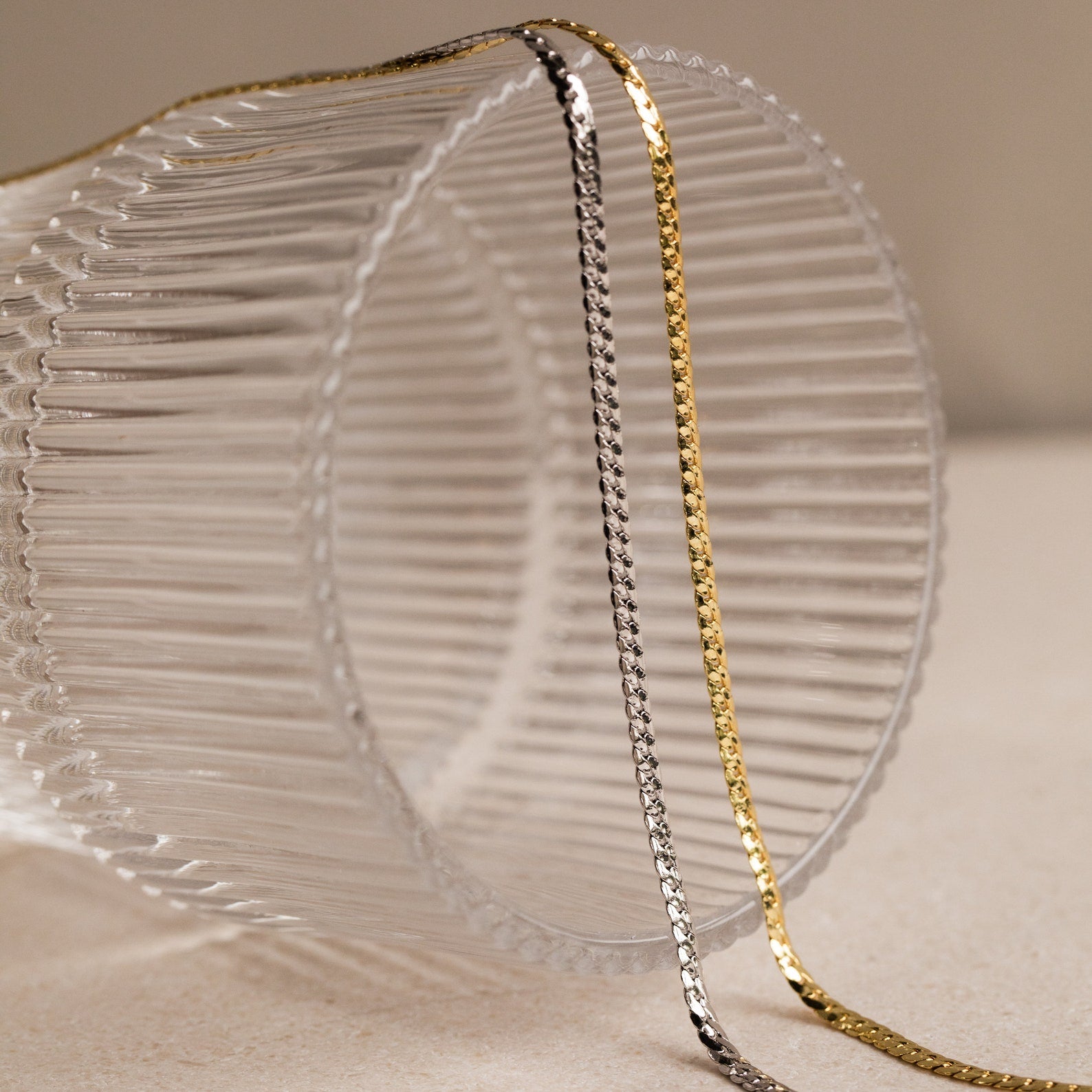 A silver Men's Snake Chain Necklace and a minimalist gold necklace rest on a ribbed clear glass set on a beige surface—stylish essentials for any men's jewelry collection.