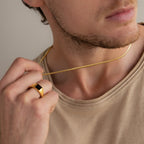 A man in a beige shirt showcases men's jewelry—a gold ring with a black stone and the Men's Snake Chain Necklace—subtly touching the minimalist necklace.