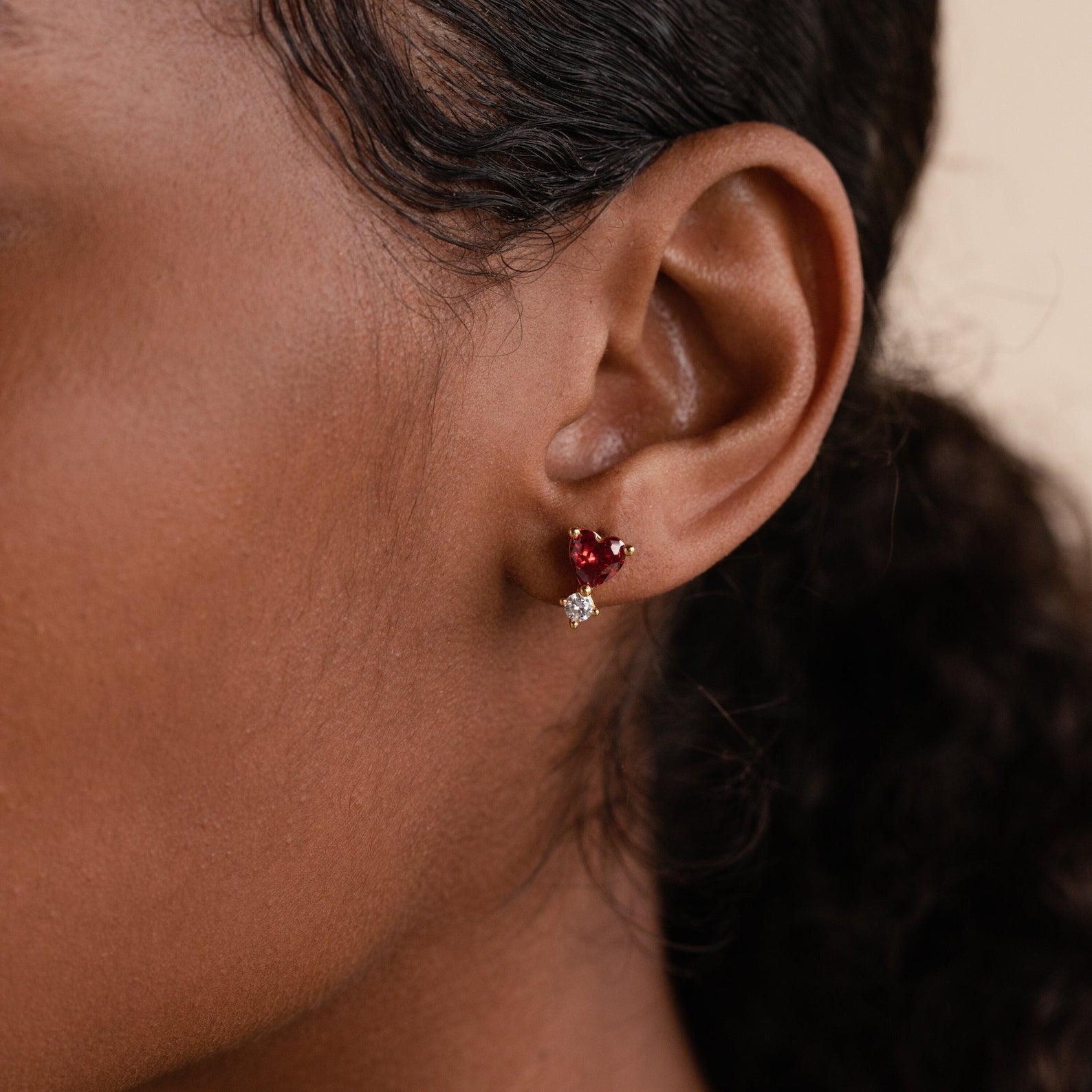 Close-up of a woman’s ear wearing Garnet Heart Studs, featuring a gold setting with a heart-shaped garnet and small clear gem—perfect for adding an elegant touch.