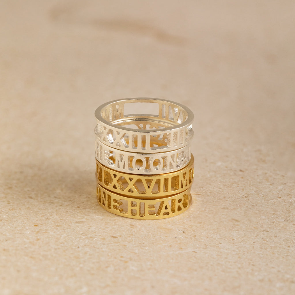 The Roman Numerals Ring in Sterling Silver features stacked silver and gold bands with cut-out numerals and lettering, creating a striking personalized jewelry piece—ideal as a milestone gift—elegantly displayed on a beige surface.