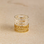 The Roman Numerals Ring in Sterling Silver features stacked silver and gold bands with cut-out numerals and lettering, creating a striking personalized jewelry piece—ideal as a milestone gift—elegantly displayed on a beige surface.