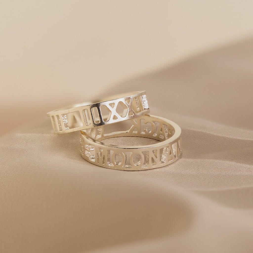 A pair of Roman Numerals Rings in sterling silver with timeless engravings rest on soft beige fabric—ideal as personalized jewelry or a milestone gift to cherish.