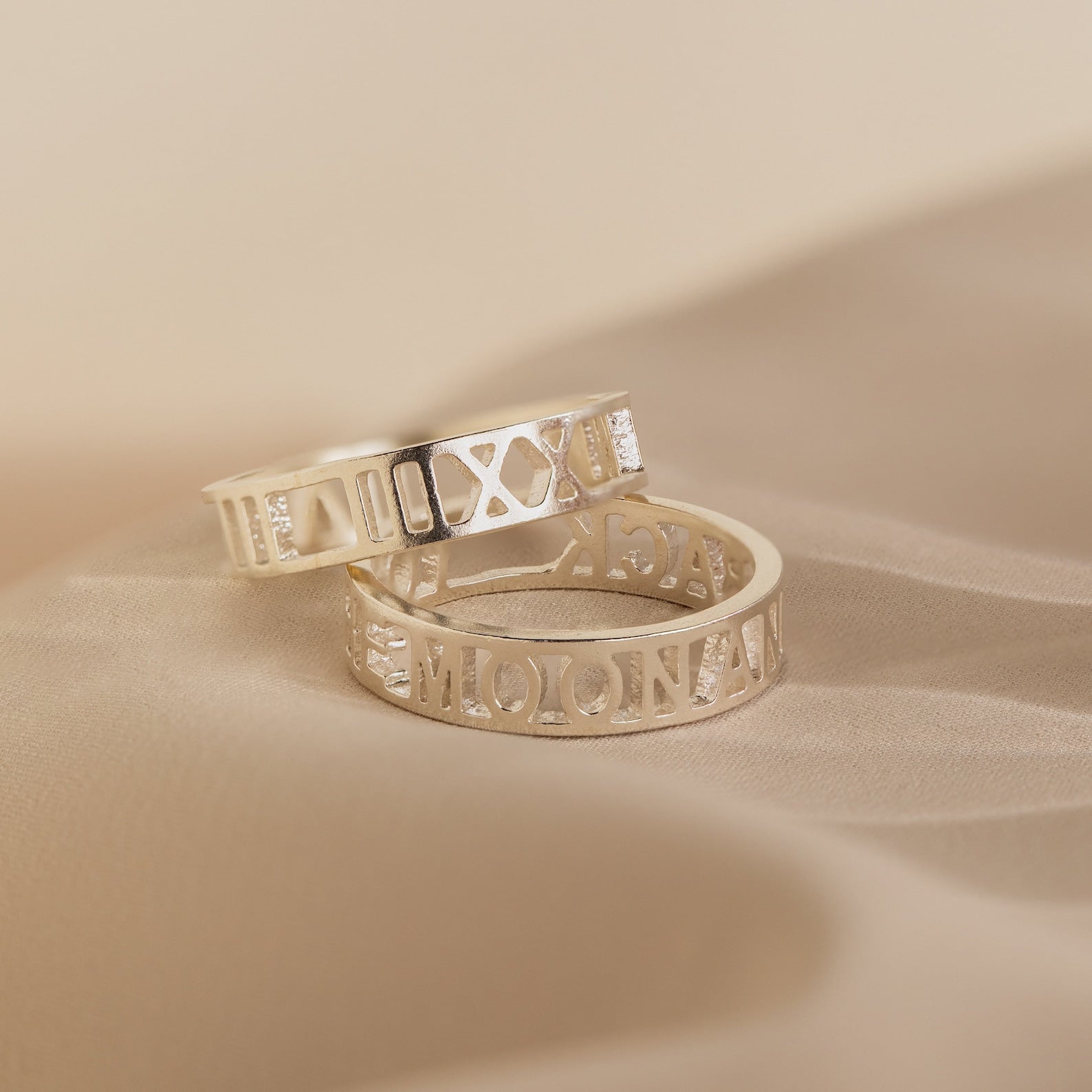 A pair of Roman Numerals Rings in sterling silver with timeless engravings rest on soft beige fabric—ideal as personalized jewelry or a milestone gift to cherish.