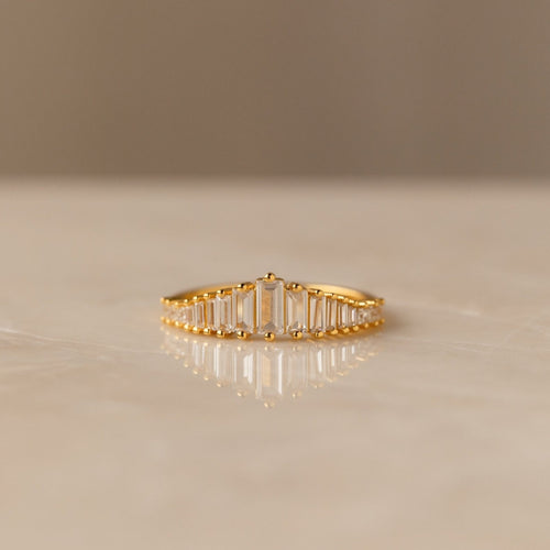 Art Deco Diamond Ring: A gold band adorned with a row of rectangular-cut clear stones, displayed elegantly on a smooth, light-colored surface—a bold statement piece.