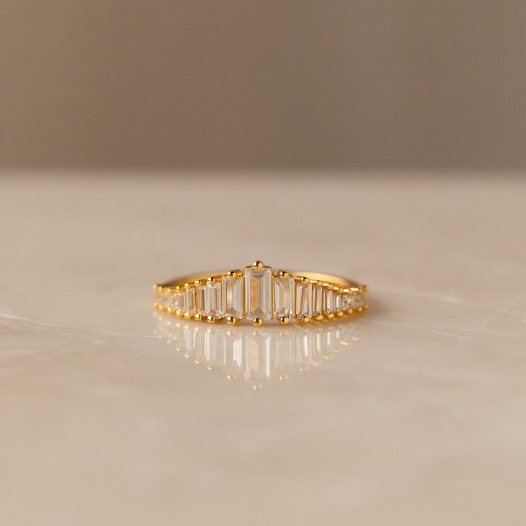 The Art Deco Huggies & Ring Set features a gold ring with a row of rectangular baguette diamonds, inspired by Art Deco jewelry, shown on a smooth light-colored surface.