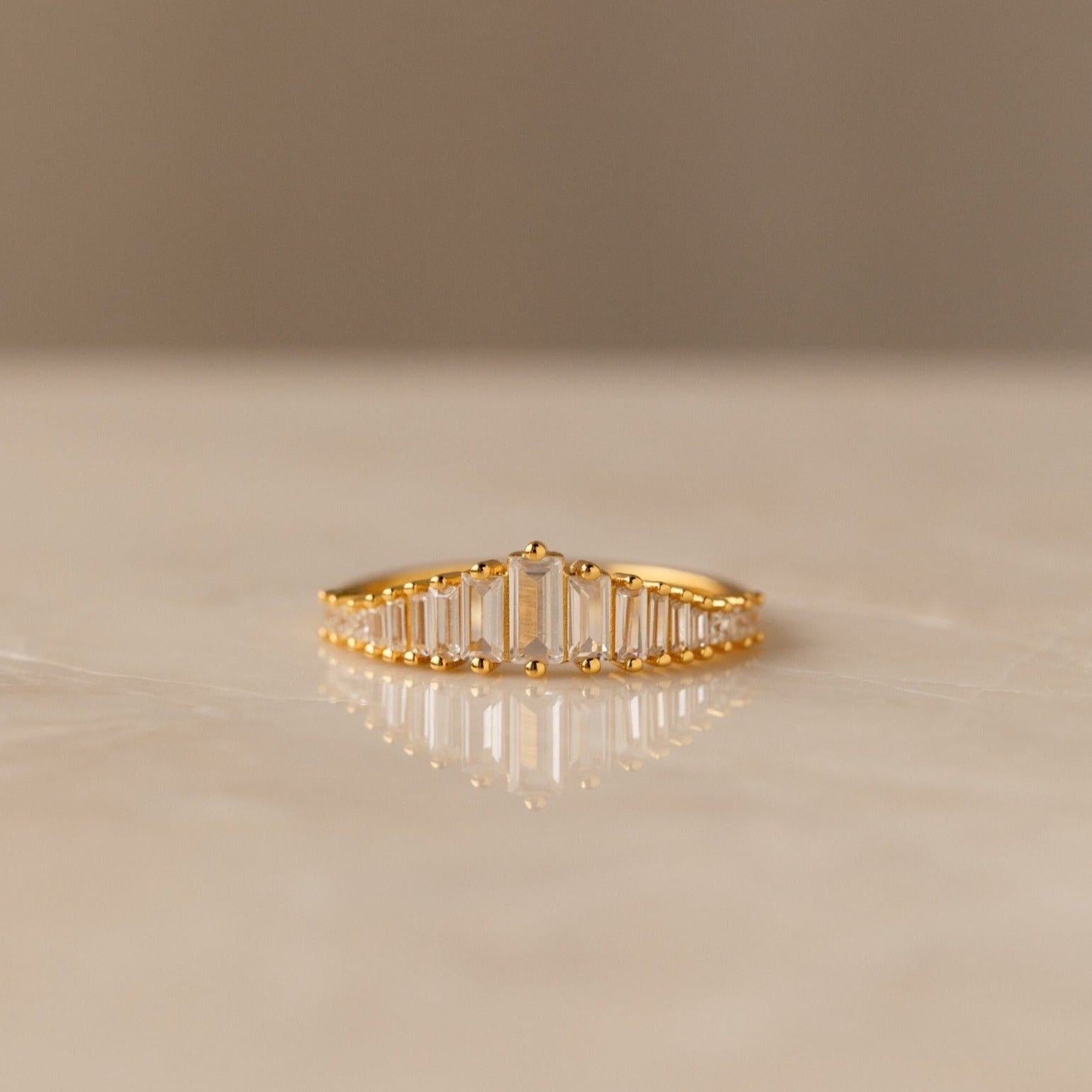 The Art Deco Huggies & Ring Set features a gold ring with a row of rectangular baguette diamonds, inspired by Art Deco jewelry, shown on a smooth light-colored surface.