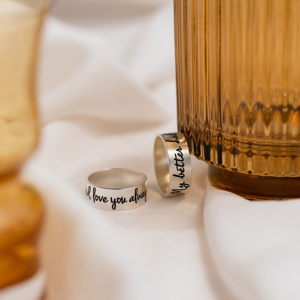 Two Thick Handwriting Band rings with engraved text lie beside a ridged amber glass on soft white fabric, perfect for fans of minimalist jewelry.