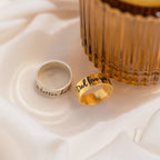 Two engraved rings, including a bold Thick Handwriting Band, lie on white fabric next to a ribbed amber glass container—adding an elegant, minimalist jewelry accent.