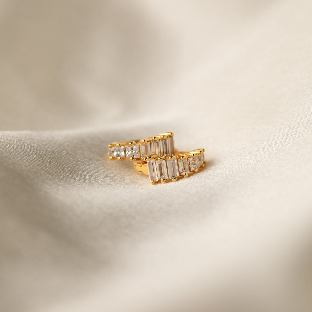 Gold Art Deco Huggie earrings resting on soft ivory fabric, showcasing the sharp contrast of geometric baguette and round stones.