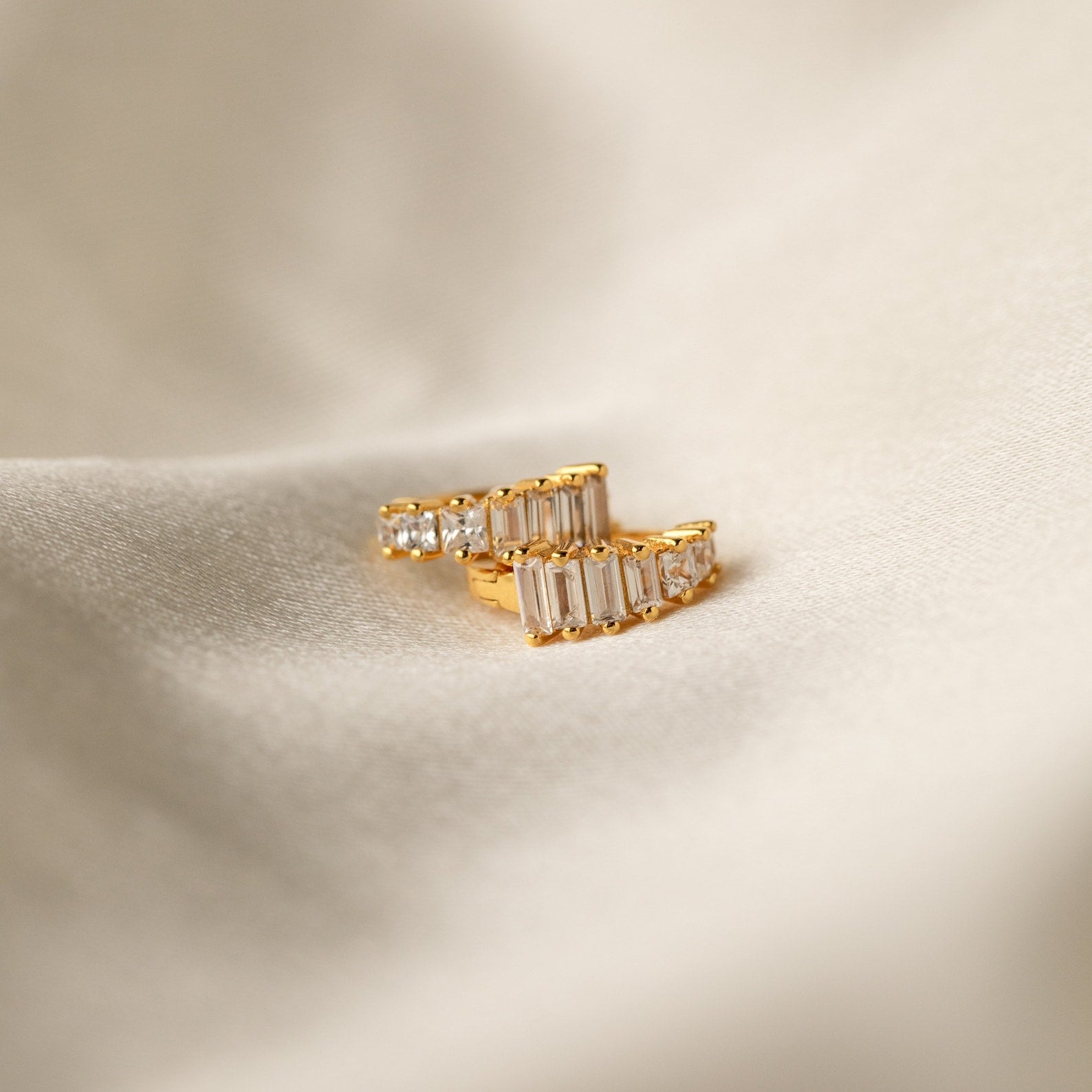 Gold Art Deco Huggie earrings resting on soft ivory fabric, showcasing the sharp contrast of geometric baguette and round stones.
