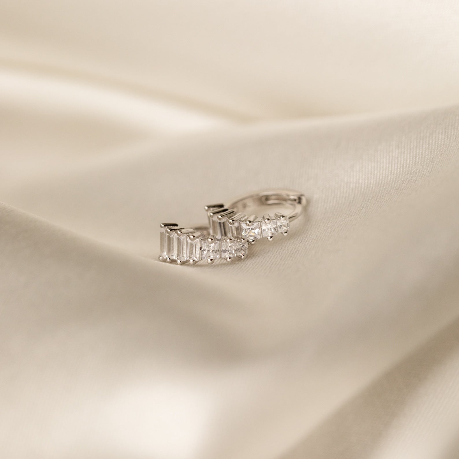 Silver version of the Art Deco Huggie earrings lying on a silky cream surface, emphasizing the clean, icy sparkle of the stones.