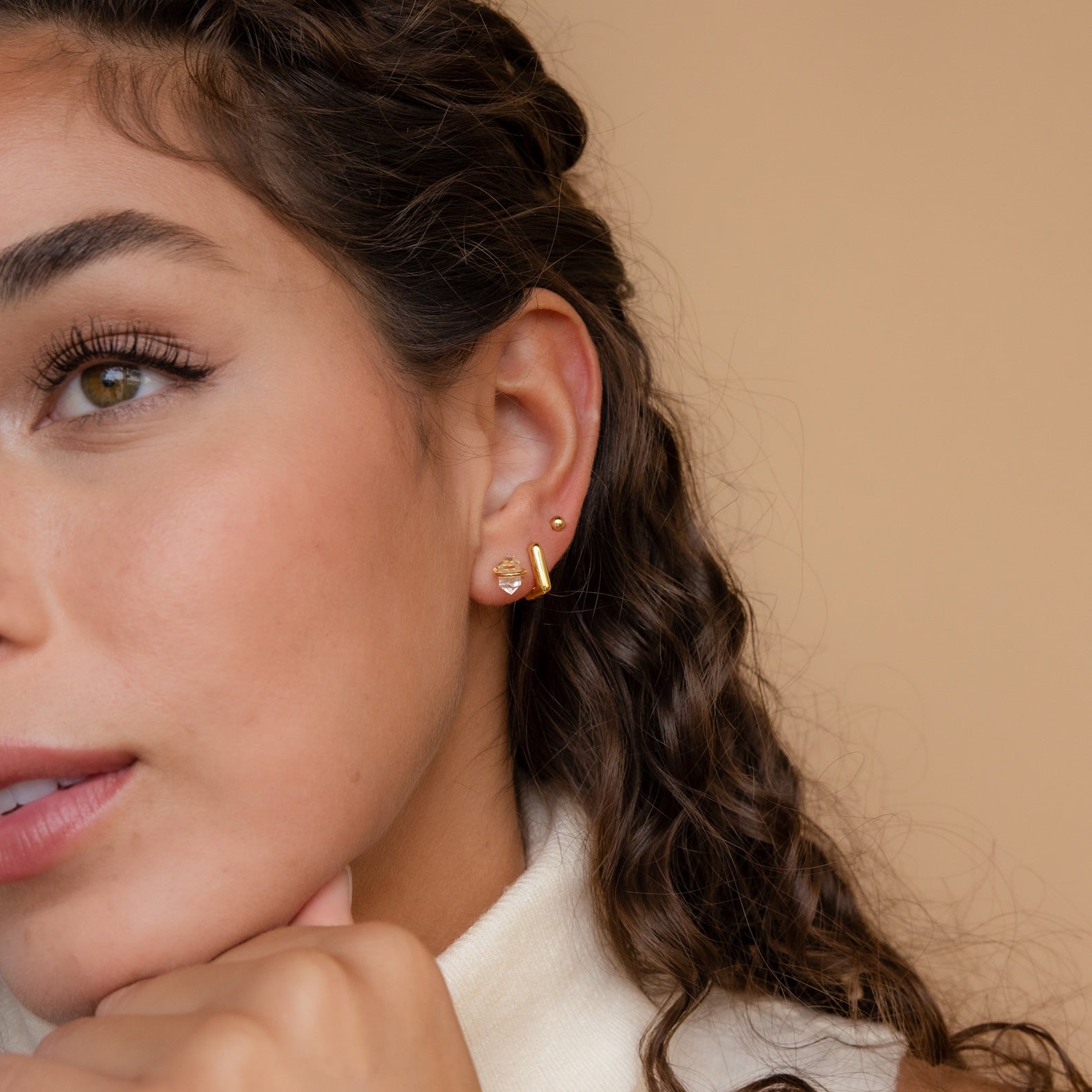 Close-up of a woman's ear wearing Herkimer Diamond Studs and multiple gold earrings, her curly hair framing the look against a neutral beige background.
