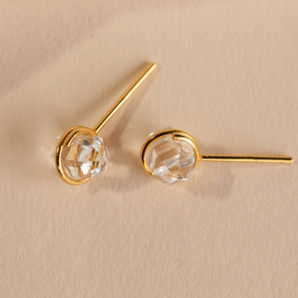 Herkimer Diamond Studs: Gold minimalist stud earrings with faceted clear Herkimer diamond settings, reminiscent of raw crystal, displayed on a beige background.