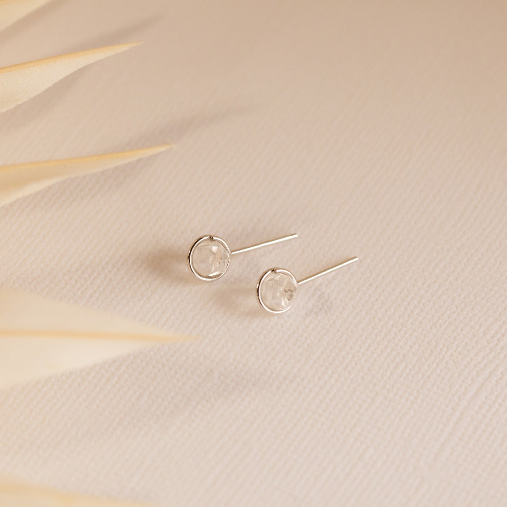 Herkimer Diamond Studs—minimalist earrings with clear stones—rest on a textured beige surface, soft leaves in the foreground.