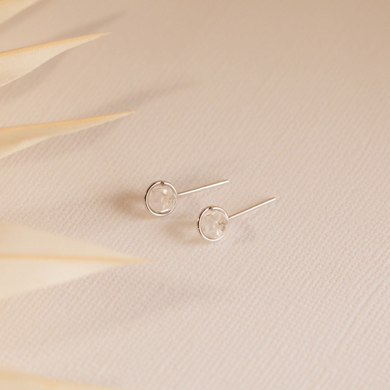 Herkimer Diamond Studs—minimalist earrings with clear stones—rest on a textured beige surface, soft leaves in the foreground.
