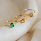 Three gold rings with emerald-cut green, peach, and blue stones are displayed on a light textured surface; one is the Citrine Emerald Ring, perfect for those with a November birthstone.