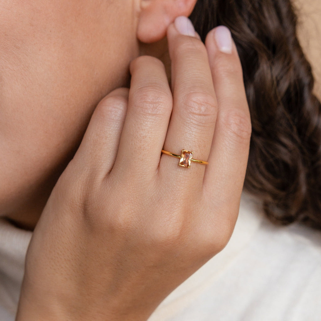 A hand wearing an Emerald Cut Solitaire Ring, showcasing a rectangular gemstone in gold, is held close to the face.