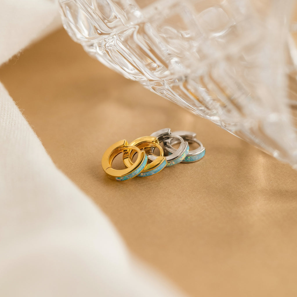 Two pairs of Ocean Opal Inlay Huggies in gold and silver rest on a tan surface beside a clear textured glass object—perfect for adding elegant charm to your wedding jewelry collection.