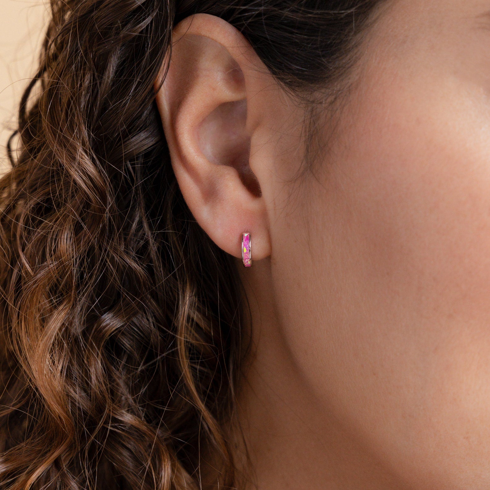 Close-up of a woman's ear wearing Magenta Opal Inlay Huggies—a small pink hoop earring styled with her curly brown hair visible.