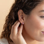 A woman with curly hair touches her ear, revealing Lavender Opal Inlay Huggies—small pink hoops that shimmer with a hint of purple opal in the light.