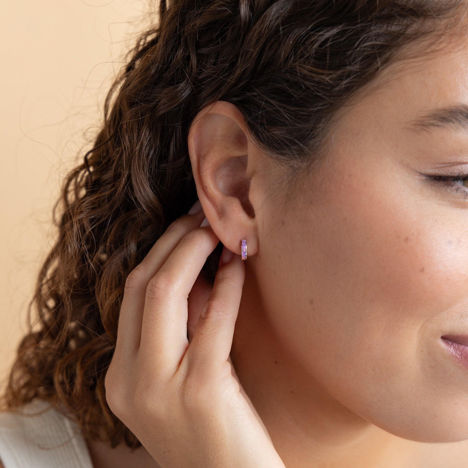 A woman with curly hair touches her ear, revealing Lavender Opal Inlay Huggies—small pink hoops that shimmer with a hint of purple opal in the light.