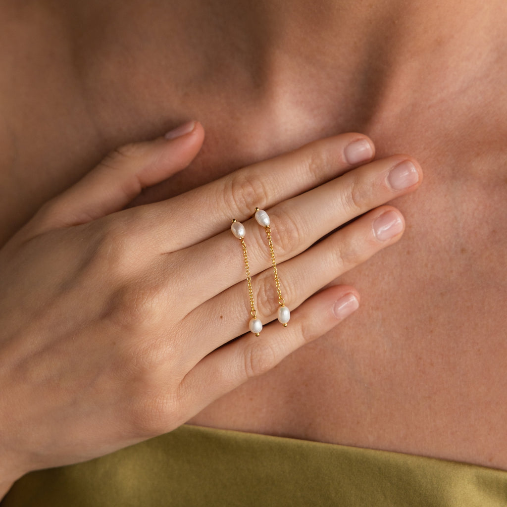 Hand with natural nails and gold rings linked by chains with pearls rests on a bare chest—beautifully complemented by our Pearl Drop Chain Earrings for an effortlessly elegant, minimalist look.