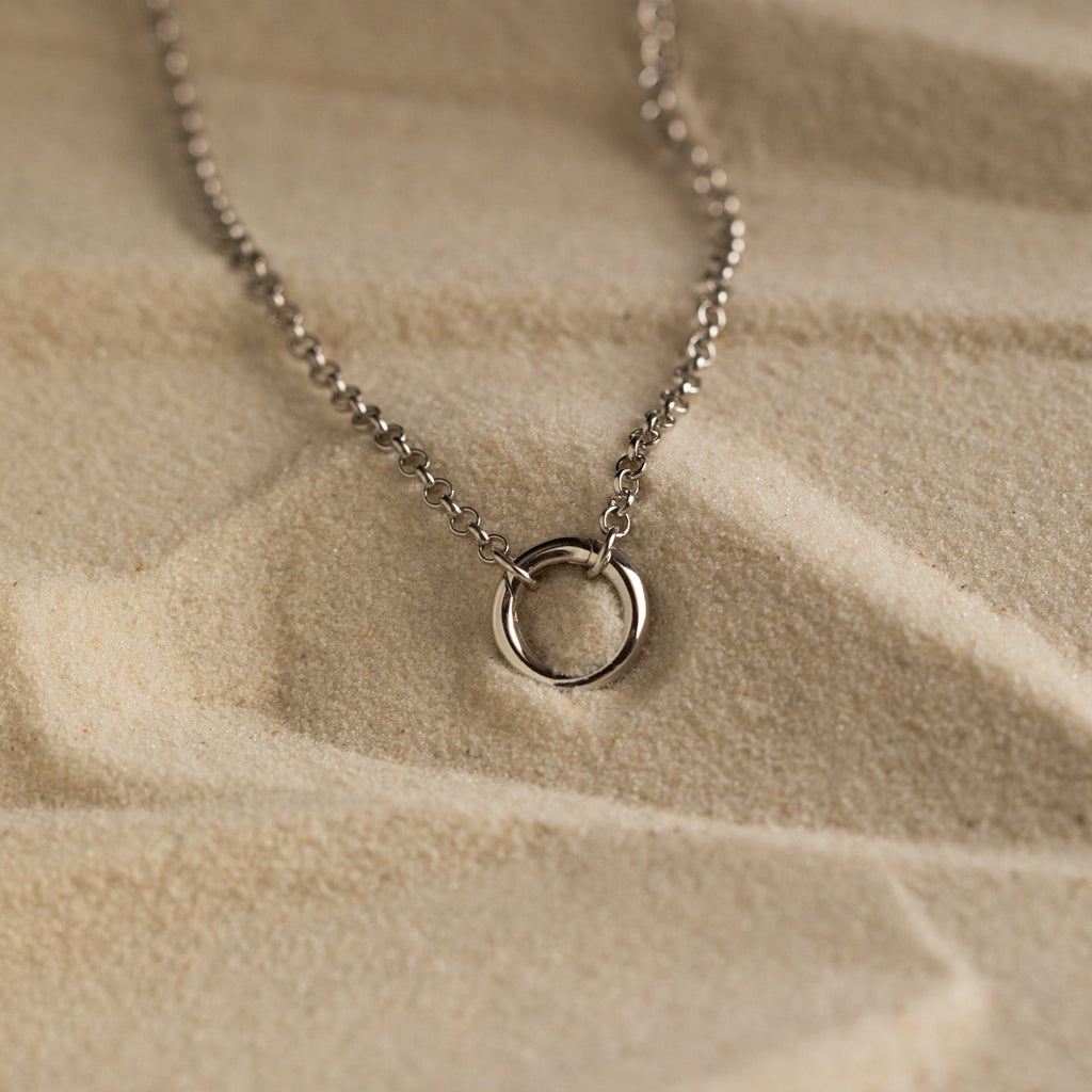 Minimalist silver necklace with a small open circle pendant on a fine chain, displayed on textured sand background.
