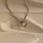 Minimalist silver necklace with a small open circle pendant on a fine chain, displayed on textured sand background.
