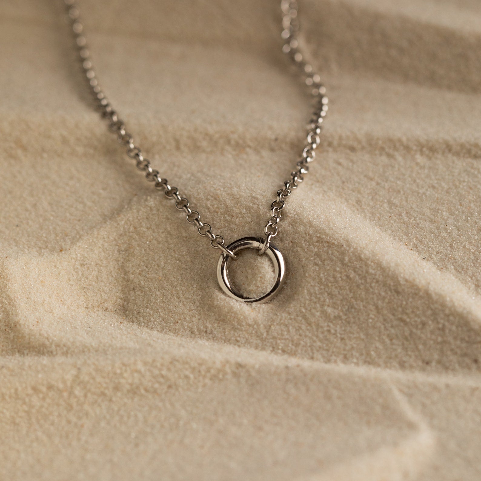 Minimalist silver necklace with a small open circle pendant on a fine chain, displayed on textured sand background.
