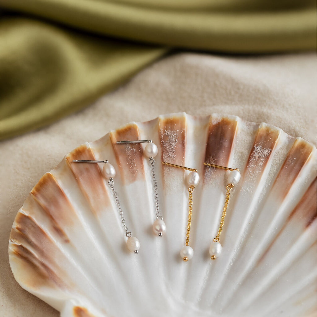 Two pairs of Pearl Drop Chain Earrings, in silver and gold, are displayed on a scallop shell with soft fabric behind them, creating a refined and minimalist presentation.
