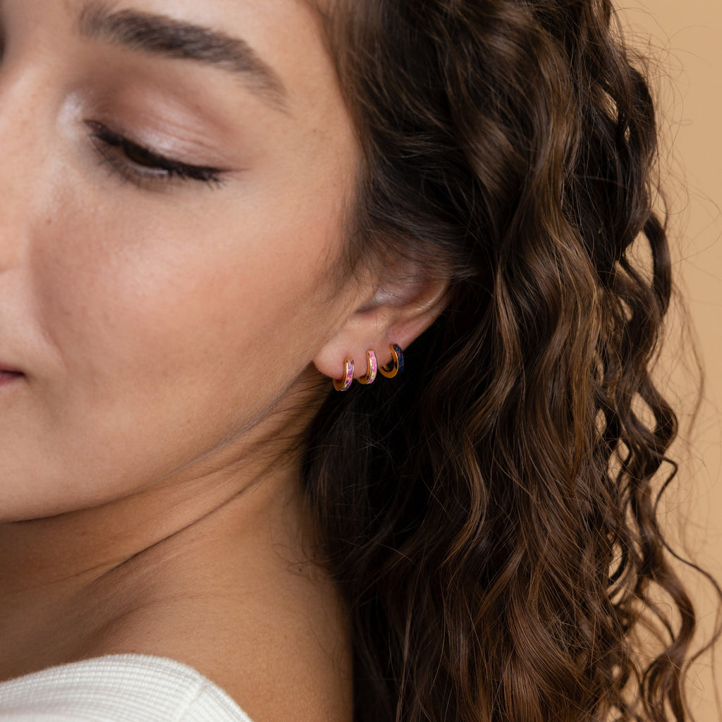 A woman with wavy brown hair wears three small, colorful hoop earrings on her ear, including minimalist hoops and the Lavender Opal Inlay Huggies.