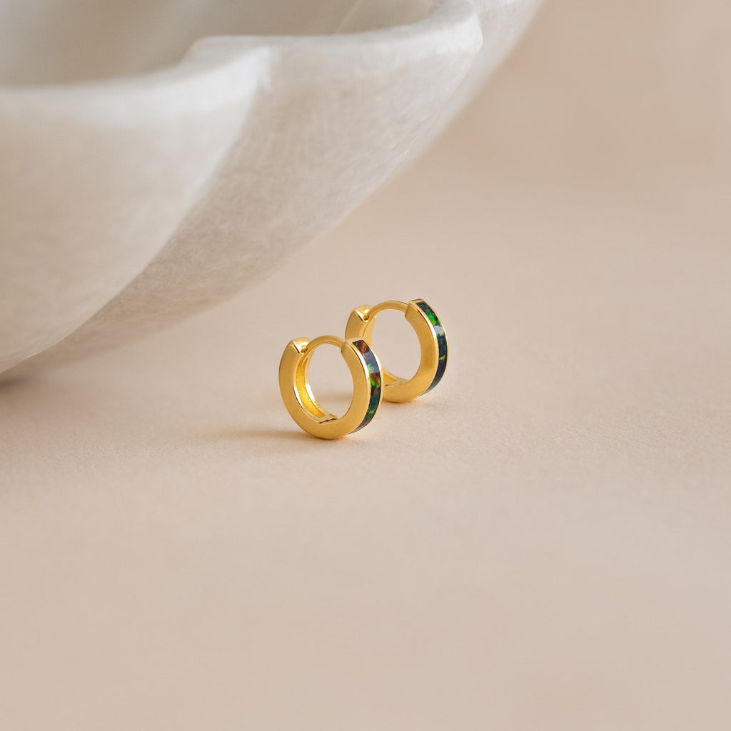 Small gold hoop earrings with a green accent—our Black Opal Inlay Huggies—are displayed on a light beige surface near a marble dish.